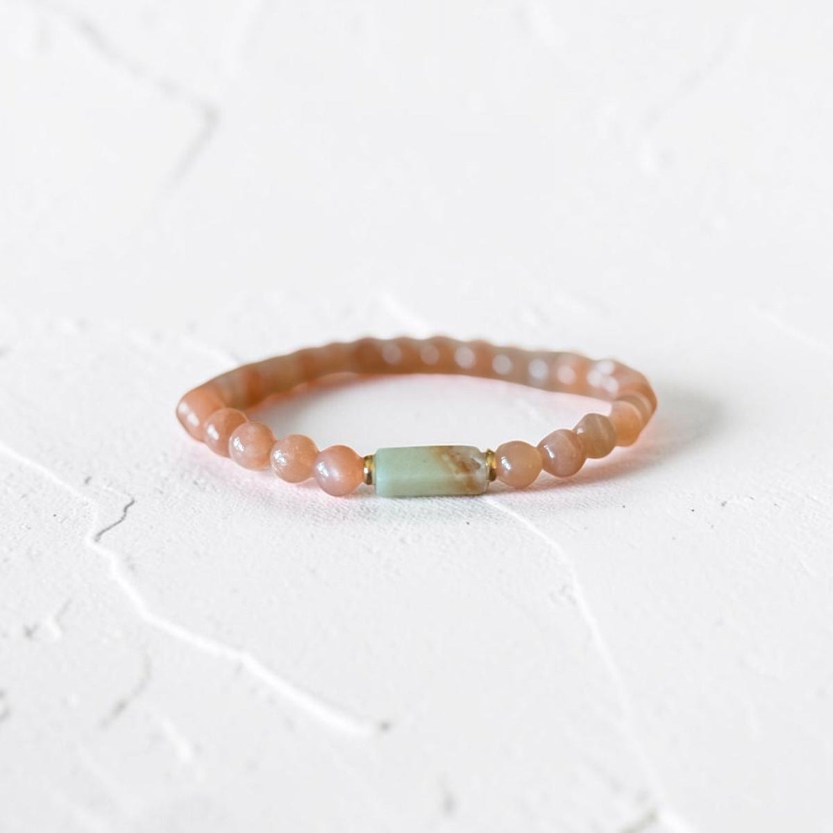 A beaded stretch bracelet featuring peachy-pink round beads and a central light green rectangular stone accent, displayed on a textured white surface.