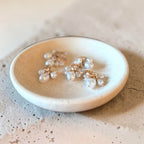 Three small floral cluster earrings with clear pearl-like beads and gold-tone settings resting in a shallow round ceramic dish on a textured beige surface.
