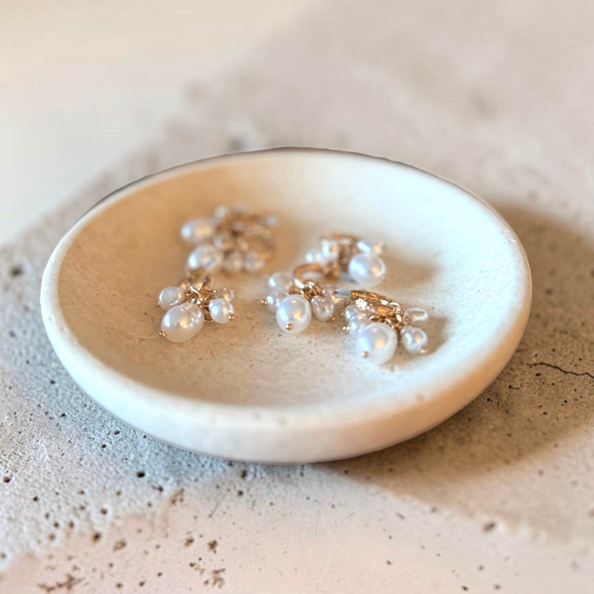 Three small floral cluster earrings with clear pearl-like beads and gold-tone settings resting in a shallow round ceramic dish on a textured beige surface.