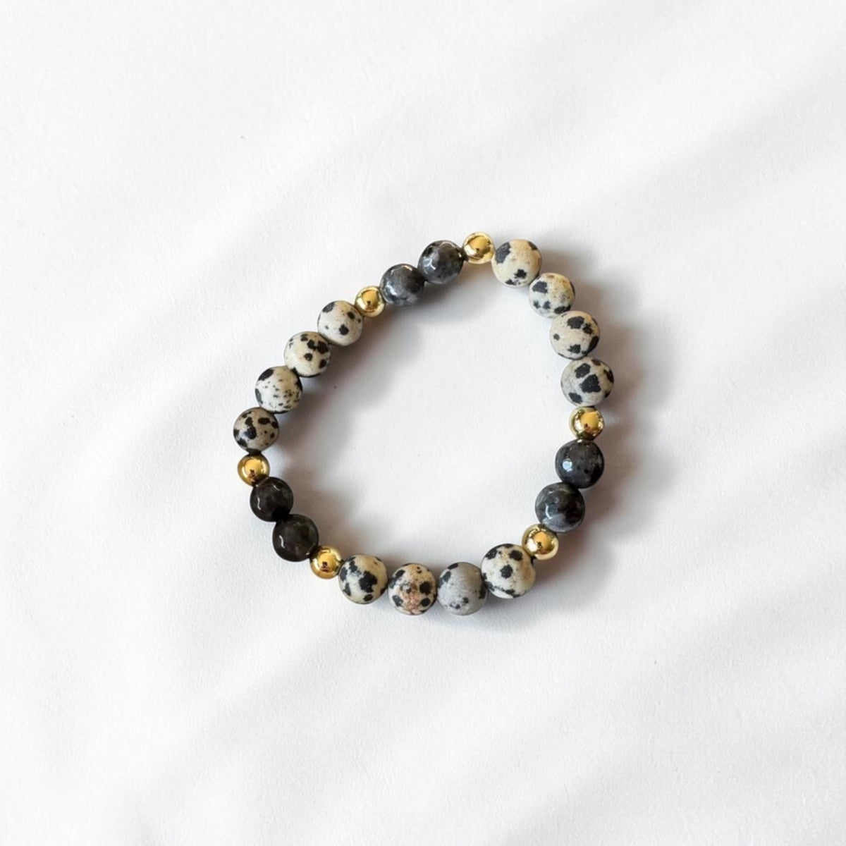 A beaded stretch bracelet featuring white and black spotted dogstone beads, dark gray beads, and small polished gold spacer beads, displayed on a white fabric background.