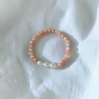 A peach-colored beaded bracelet with three white teardrop-shaped freshwater pearls and a small gold clasp, resting on a white fabric background.