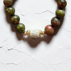 A beaded bracelet with round green and pinkish stone beads, featuring a central textured white bead flanked by small gold spacer rings, shown on a white textured background.