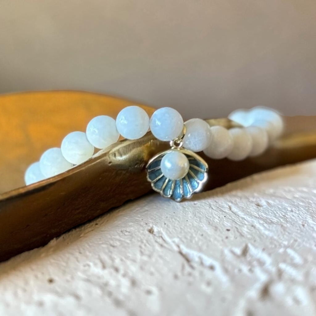 A beaded stretch bracelet with smooth white oval beads and a small shell-shaped charm featuring a white pearl center and blue-gray detailing, resting on a wooden surface.