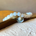 A beaded stretch bracelet with smooth white oval beads and a small shell-shaped charm featuring a white pearl center and blue-gray detailing, resting on a wooden surface.