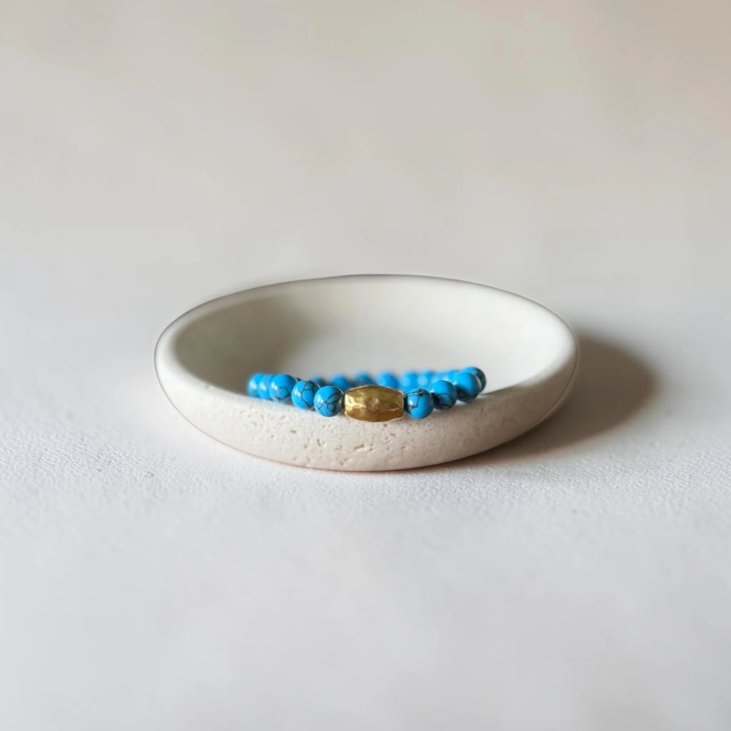 A beaded bracelet with bright blue beads and a central gold-toned oval charm, displayed inside a small round off-white ceramic dish on a light neutral background.