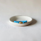 A beaded bracelet with bright blue beads and a central gold-toned oval charm, displayed inside a small round off-white ceramic dish on a light neutral background.