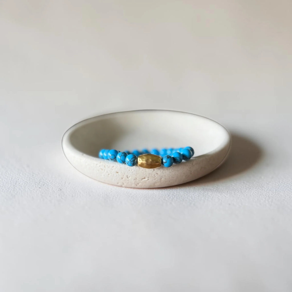 A beaded bracelet with bright blue beads and a central gold-toned oval charm, displayed inside a small round off-white ceramic dish on a light neutral background.