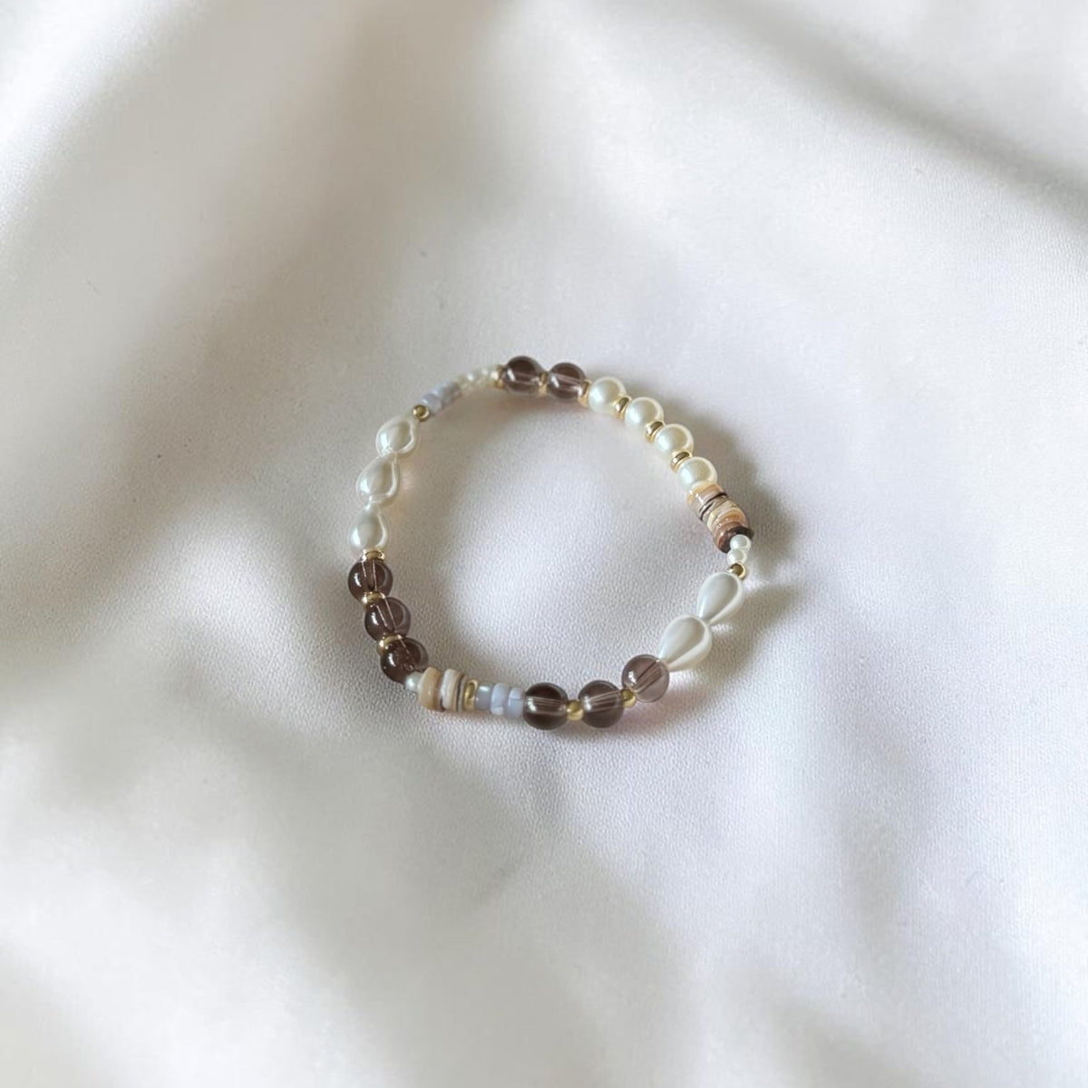 A beaded stretch bracelet featuring white pearls, brown beads, and small iridescent accents, displayed on a soft, draped white fabric background.