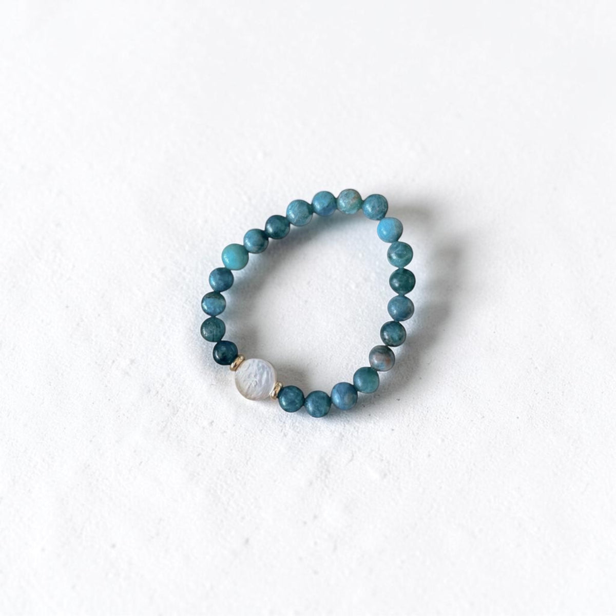 A beaded stretch bracelet featuring round teal-blue and dark blue beads, accented by a single white moon-phase bead with a gold-toned rim, displayed on a textured white background.