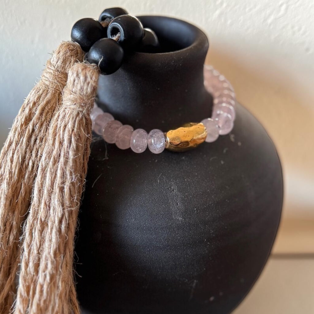 A beaded bracelet with pale pink translucent beads, a textured gold-toned accent bead, and black round beads, draped around a matte black ceramic vase. A long tan jute tassel hangs from the left side.