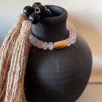 A beaded bracelet with pale pink translucent beads, a textured gold-toned accent bead, and black round beads, draped around a matte black ceramic vase. A long tan jute tassel hangs from the left side.