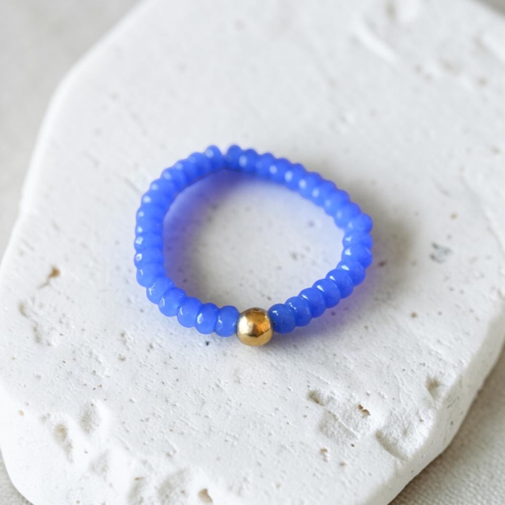 A beaded stretch bracelet in vibrant blue with a single gold accent bead, displayed on a textured white stone surface.