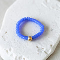 A beaded stretch bracelet in vibrant blue with a single gold accent bead, displayed on a textured white stone surface.