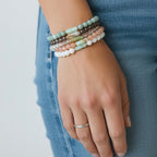 A close-up of a person's hand wearing a stack of five beaded stretch bracelets in pastel colors, paired with a silver band ring and blue jeans.