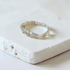 A delicate beaded bracelet with iridescent pearl-like beads and small gold-toned accents, resting on a textured white stone slab against a soft white background.