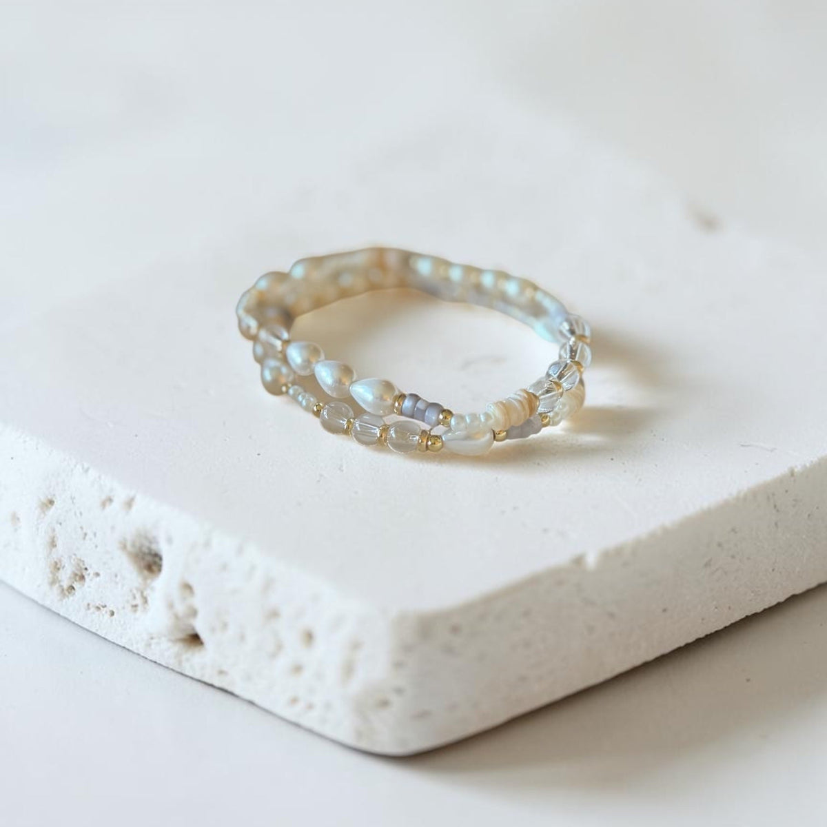 A delicate beaded bracelet with iridescent pearl-like beads and small gold-toned accents, resting on a textured white stone slab against a soft white background.