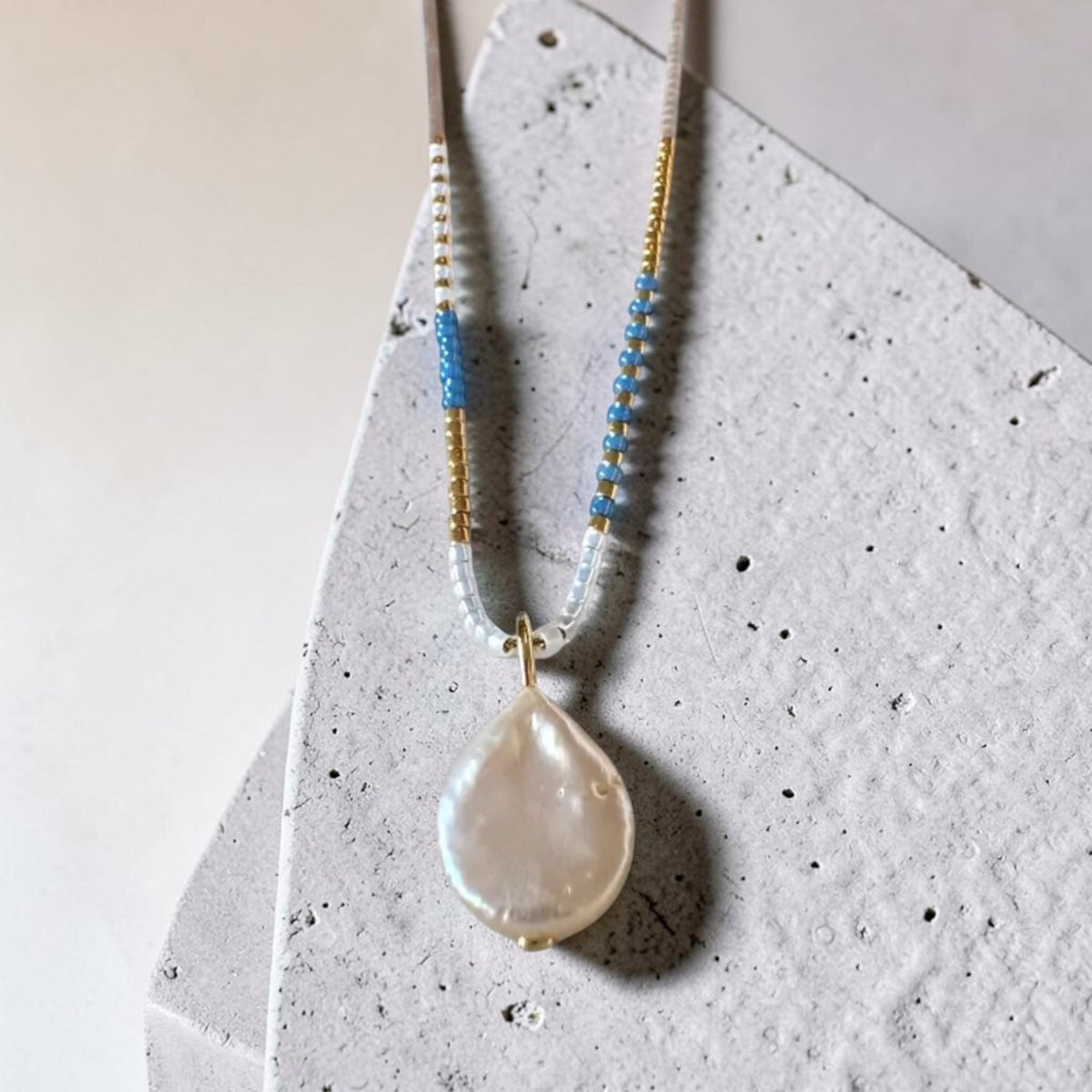 A beaded necklace with a large, irregular teardrop-shaped pearl pendant, displayed on a textured light gray stone slab. The necklace features white and light blue beads with small gold spacer beads and a gold-toned clasp.