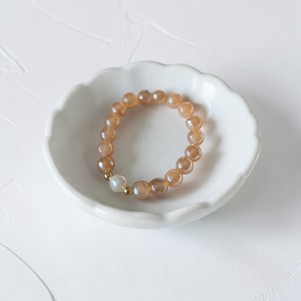 A beaded stretch bracelet with peachy-brown translucent round beads, a small white pearl-like bead, and gold spacer accents, displayed in a small white ceramic dish on a light textured background.