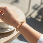 A close-up lifestyle shot of a person's wrist wearing a beaded bracelet with white pearls and a central gold-toned bead, set against a warm, sunlit cafe table background.