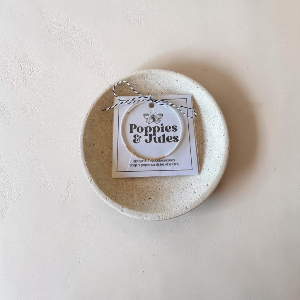 A small round ceramic dish in a speckled cream-beige glaze, photographed from above on a textured off-white surface. A paper tag reading 'Poppies & Jades' is tied to the rim with twine.