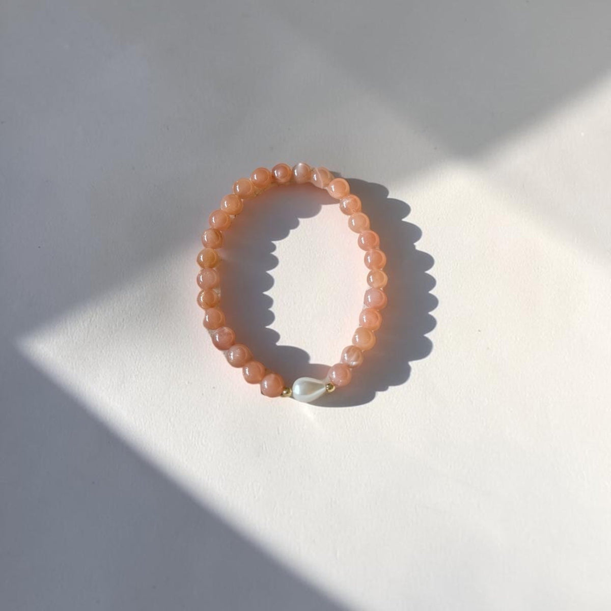 A peach-orange beaded stretch bracelet with a small white shell accent, photographed on a white surface with strong natural sunlight and shadows.