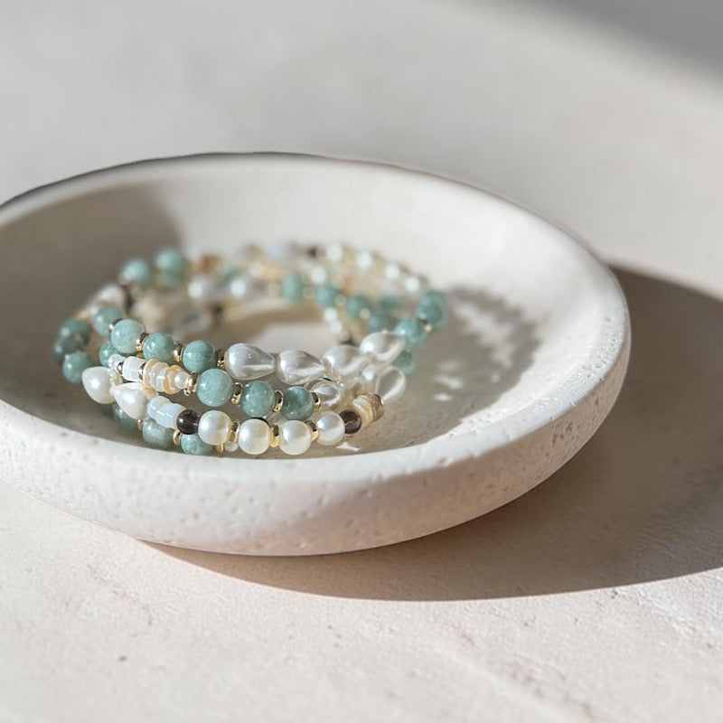 A stack of beaded stretch bracelets in light blue-green, white, and beige tones, displayed inside a small speckled ceramic dish on a sunlit neutral surface.