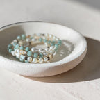 A stack of beaded stretch bracelets in light blue-green, white, and beige tones, displayed inside a small speckled ceramic dish on a sunlit neutral surface.