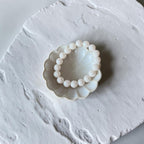 A beaded bracelet made of 10mm round, off-white beads arranged in a circle, resting on a small scalloped ceramic dish over a textured white surface.