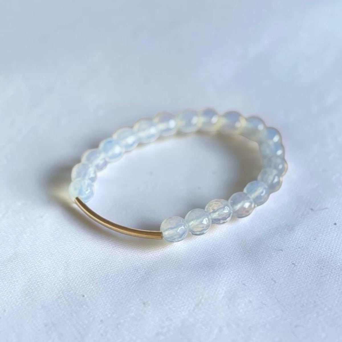 A beaded stretch ring featuring translucent white glass beads and a polished gold-toned metal bar, resting on a white textured fabric background.