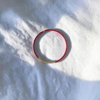 A delicate red beaded bracelet with a small gold bead accent on one end, photographed on a white textured fabric background with soft dappled sunlight and shadows.