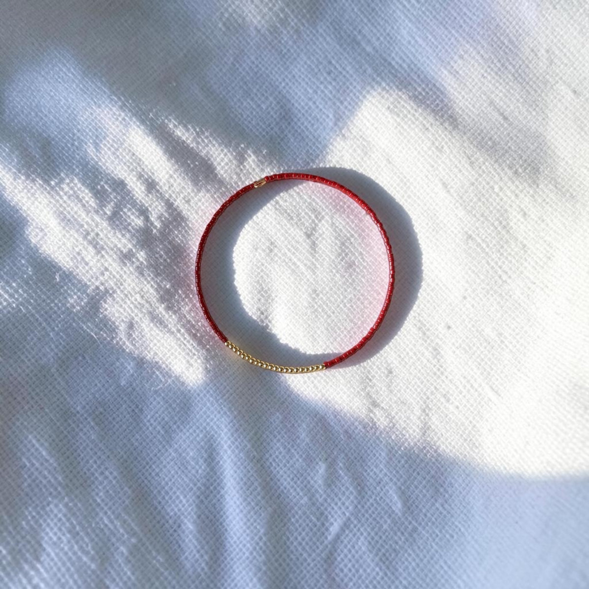 A delicate red beaded bracelet with a small gold bead accent on one end, photographed on a white textured fabric background with soft dappled sunlight and shadows.