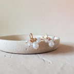 A pair of gold-toned dangle earrings with irregular white freshwater pearls, displayed on a textured beige ceramic dish against a soft beige background.
