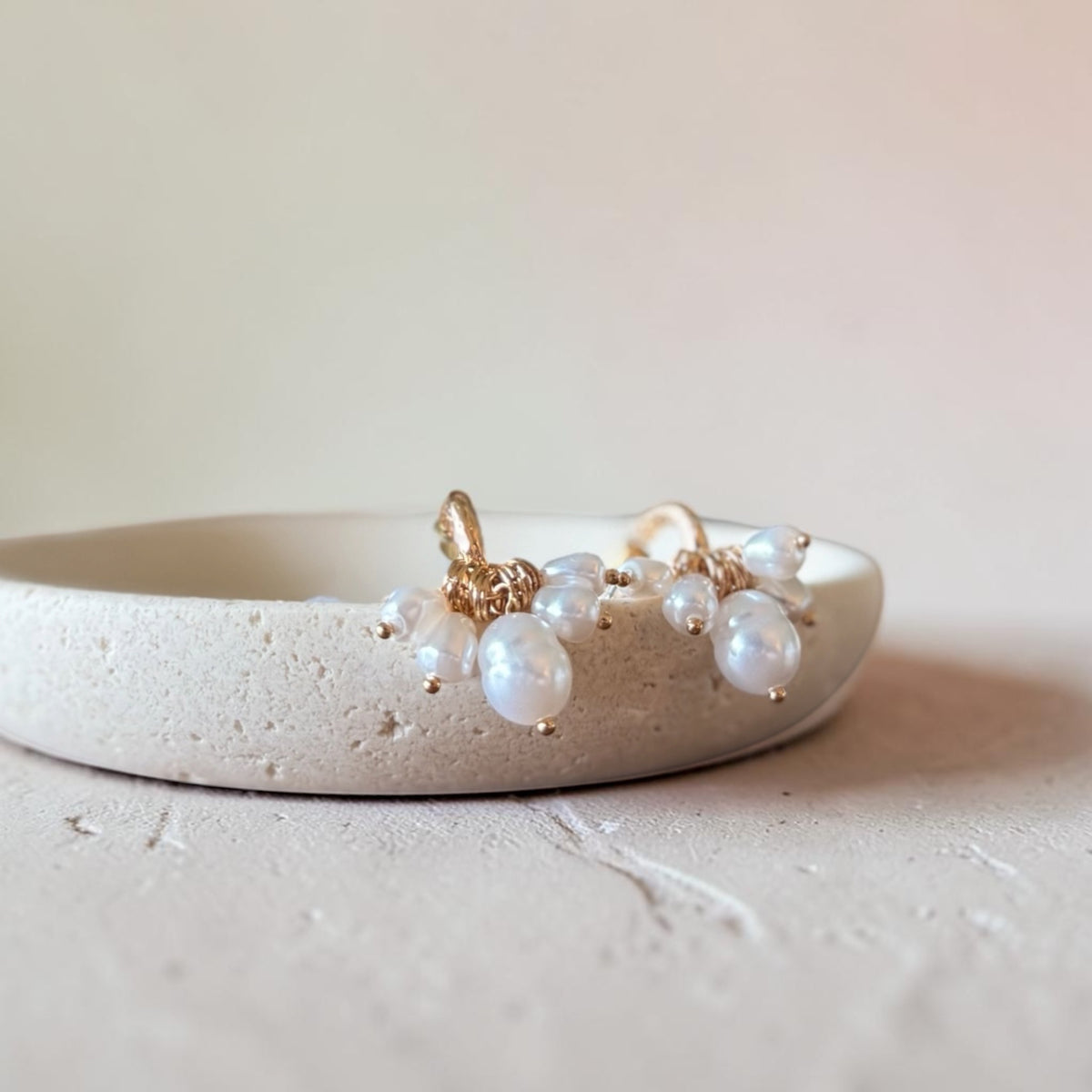 A pair of gold-toned dangle earrings with irregular white freshwater pearls, displayed on a textured beige ceramic dish against a soft beige background.