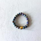A beaded stretch bracelet made of iridescent dark beads with a central gold-toned spacer bead, displayed on a textured off-white surface.