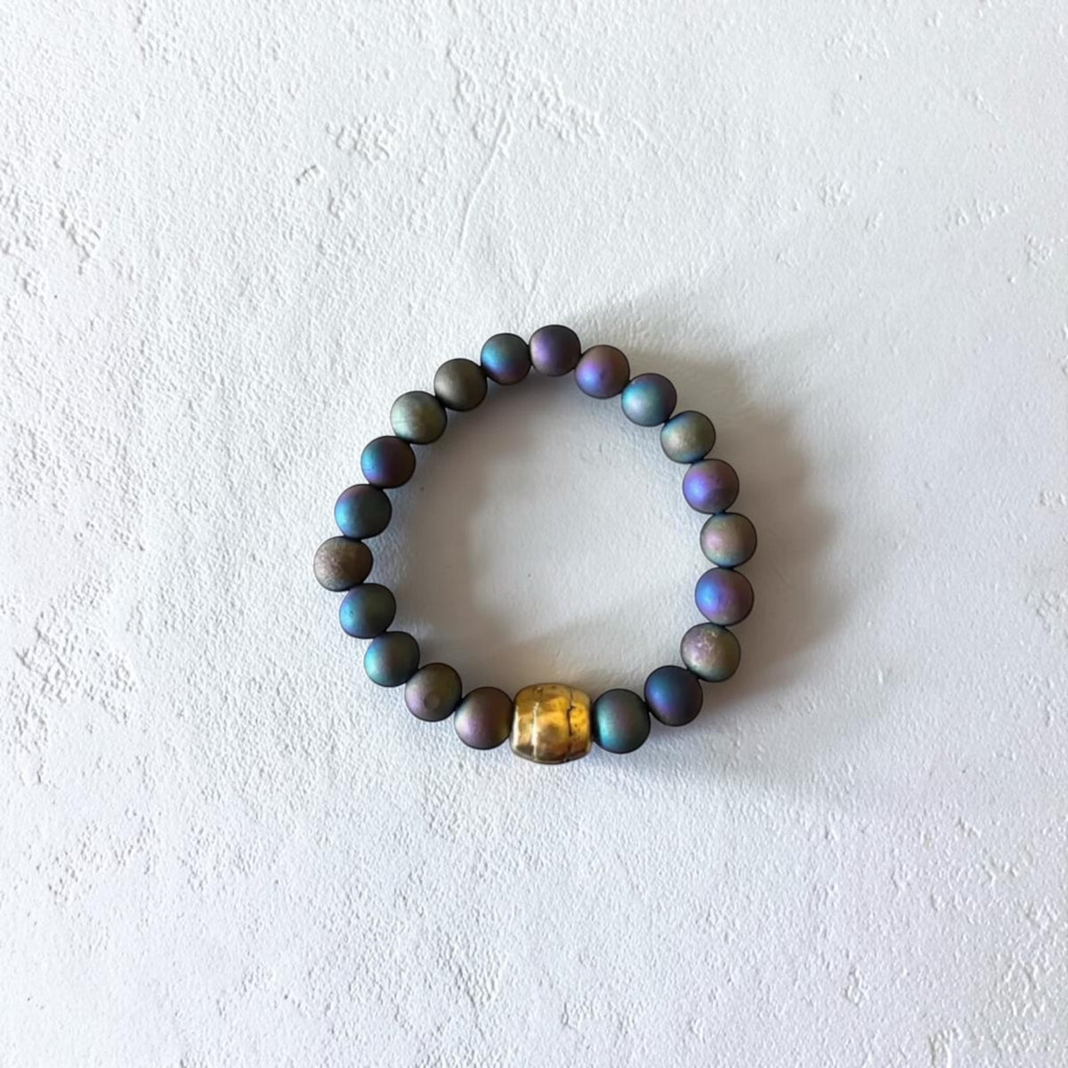 A beaded stretch bracelet made of iridescent dark beads with a central gold-toned spacer bead, displayed on a textured off-white surface.