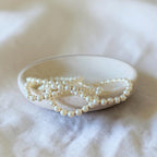A strand of cream pearl-like beads draped over a small round ceramic dish on a soft white fabric background.