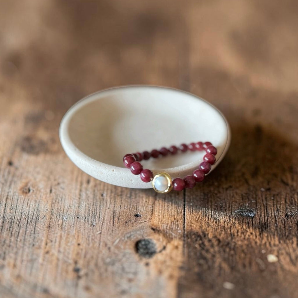 8mm Red Garnet Jade Bead Bracelet with a Gold And Pearl Accent. Accent has a round pearl with gold surrounding the pearl itself. This bracelet is sitting in a dish.