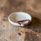 8mm Red Garnet Jade Bead Bracelet with a Gold And Pearl Accent. Accent has a round pearl with gold surrounding the pearl itself. This bracelet is sitting in a dish.