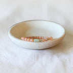 A delicate beaded bracelet with peach-pink beads and a light green central accent, displayed inside a small, round, speckled off-white ceramic dish on a soft, textured white fabric background.