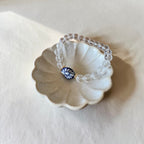 A beaded stretch bracelet made of clear faceted beads with a single blue and white floral bead and gold trim, displayed on a small scalloped ceramic dish against a light beige background.