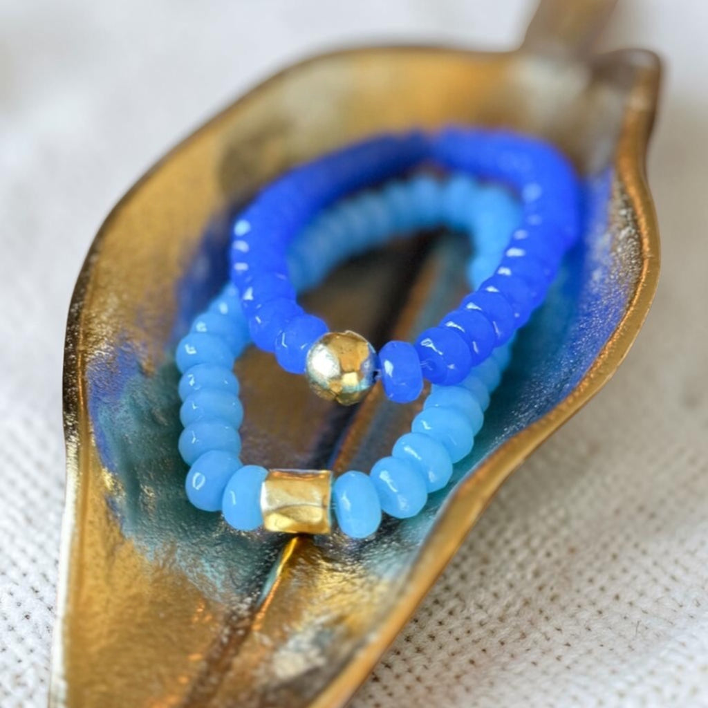 Two beaded stretch bracelets, one in royal blue and one in light sky blue, each featuring a small gold-toned faceted bead, displayed on a gold leaf-shaped dish over a white textured fabric background.