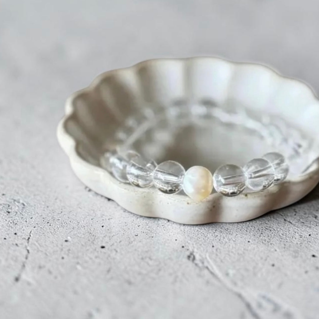A beaded bracelet with clear faceted beads and one white pearl-like bead, displayed inside a scalloped off-white ceramic dish on a textured light gray surface.