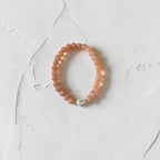 A peach-colored beaded bracelet with a small white pearl accent, displayed on a textured off-white surface.