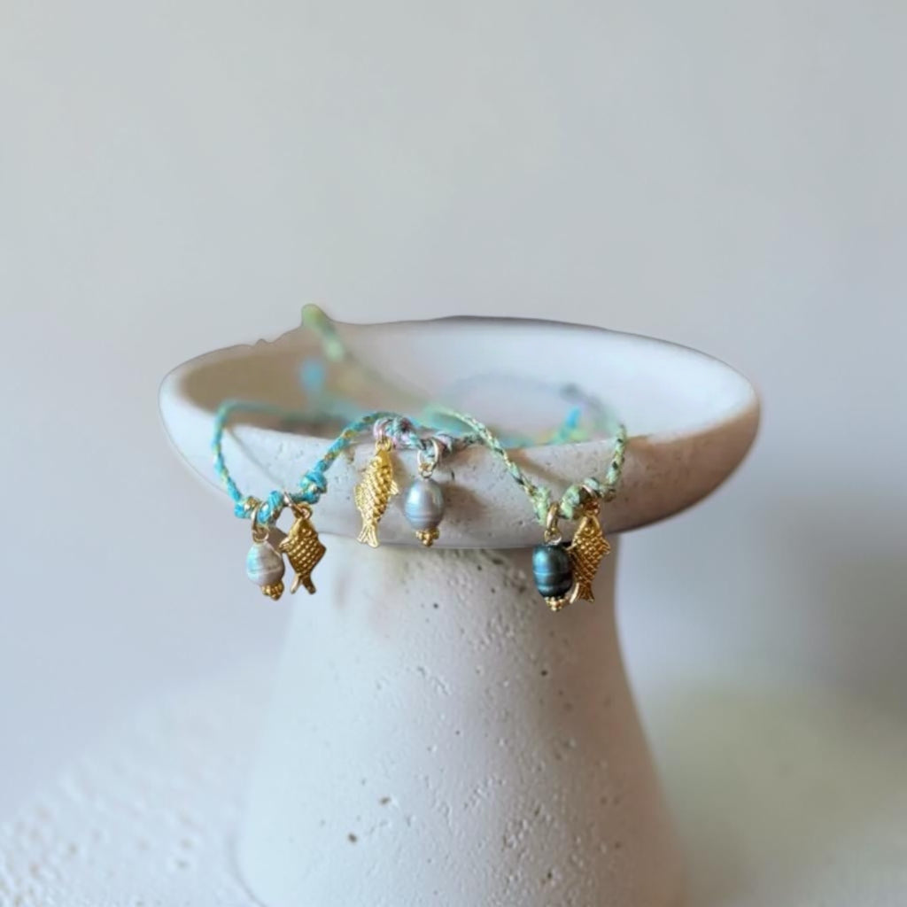 A delicate bracelet with turquoise beads, gold fish charms, and white pearl-like beads draped over a small white ceramic dish against a soft beige background.
