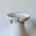 A delicate bracelet with turquoise beads, gold fish charms, and white pearl-like beads draped over a small white ceramic dish against a soft beige background.