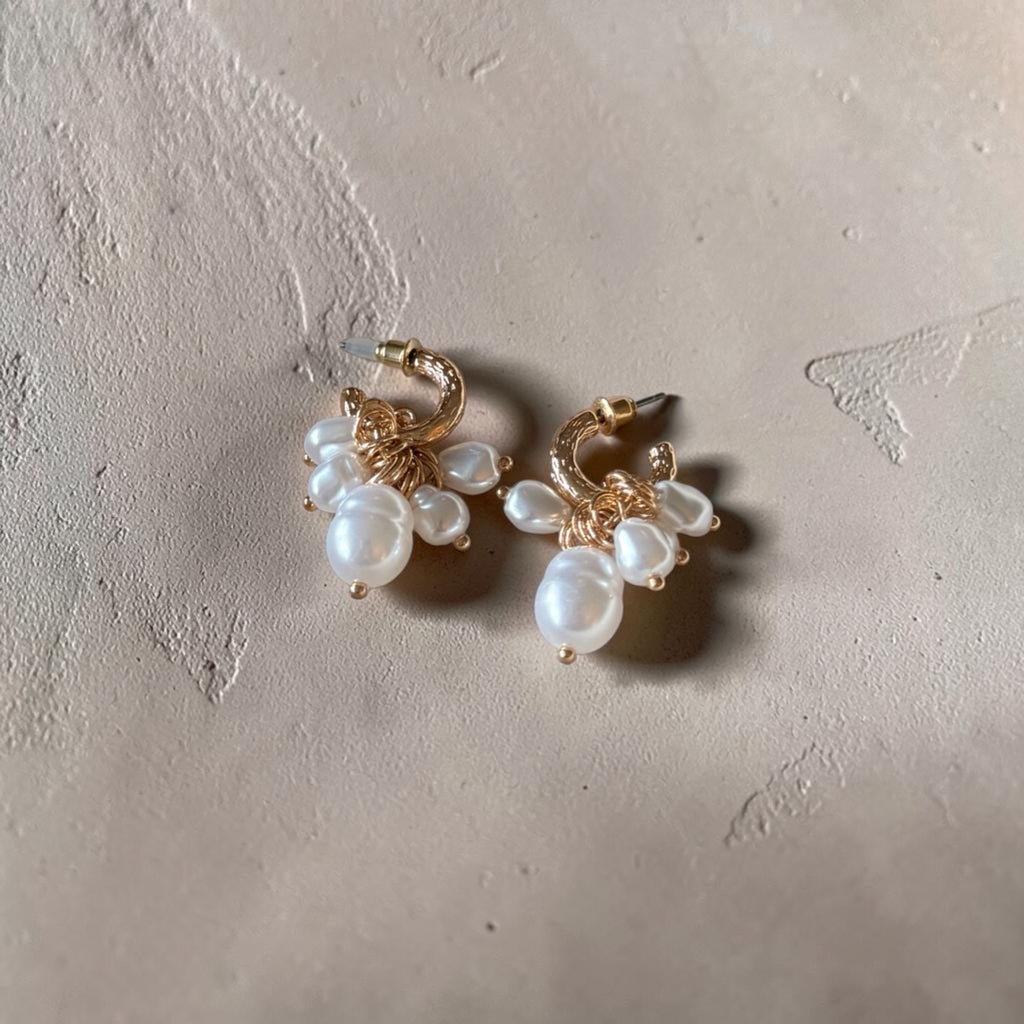 A pair of gold hoop earrings with dangling clusters of white freshwater pearls, displayed on a textured beige surface.