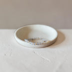 A delicate beaded stretch bracelet with iridescent, pale blue-gray beads resting inside a small, shallow, textured ceramic dish on a neutral light gray surface.