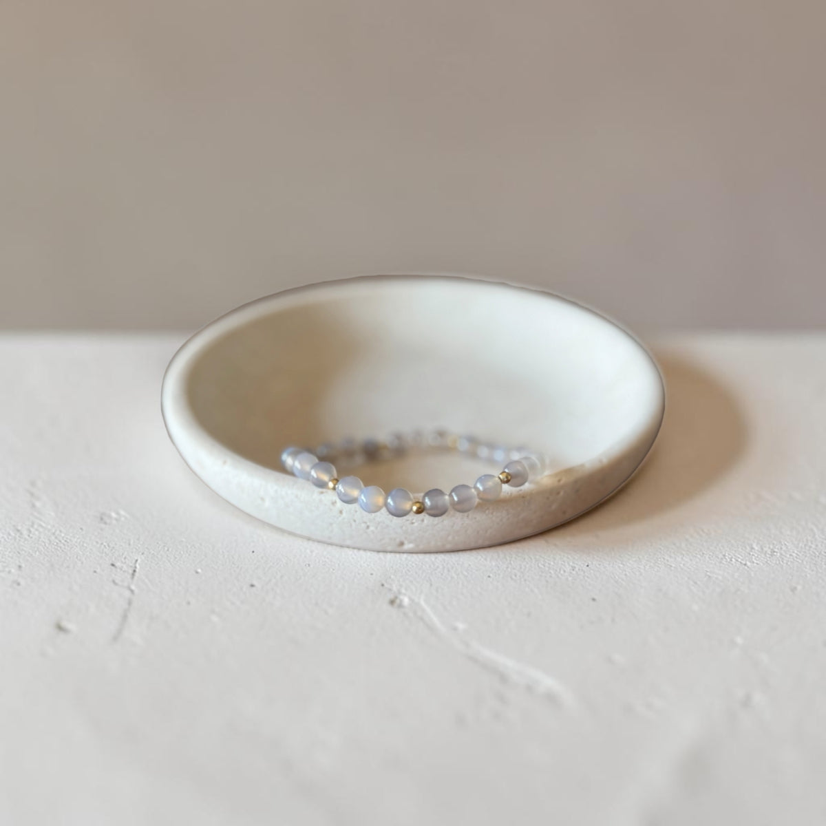 A delicate beaded stretch bracelet with iridescent, pale blue-gray beads resting inside a small, shallow, textured ceramic dish on a neutral light gray surface.