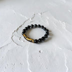 A beaded stretch bracelet featuring glossy black round beads and a curved, polished brown and black marbled accent bead, displayed on a textured white surface.