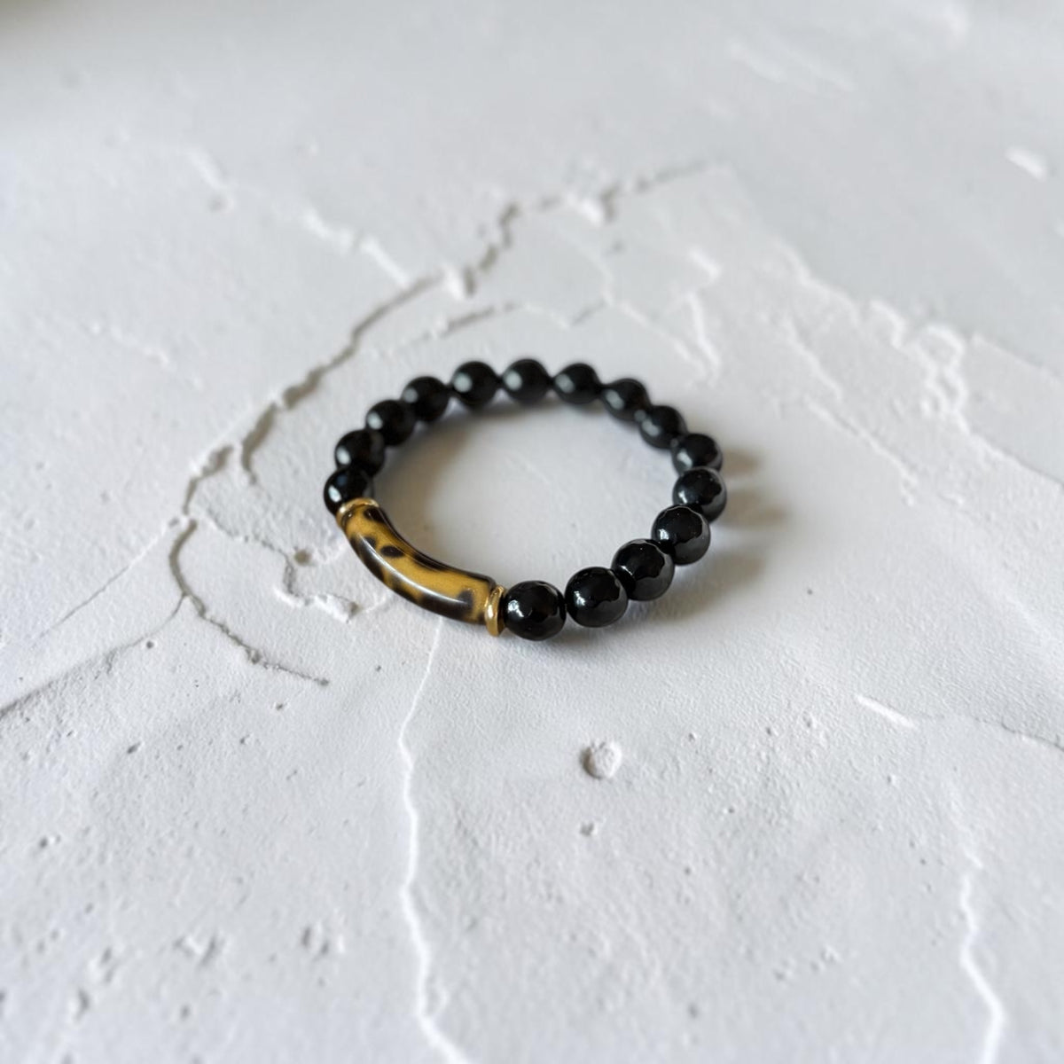 A beaded stretch bracelet featuring glossy black round beads and a curved, polished brown and black marbled accent bead, displayed on a textured white surface.
